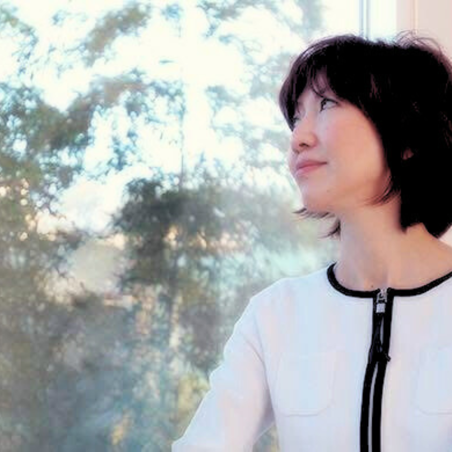 窓の外の木々と空を眺めながら立つ、姿勢改善指導者ノリコの姿